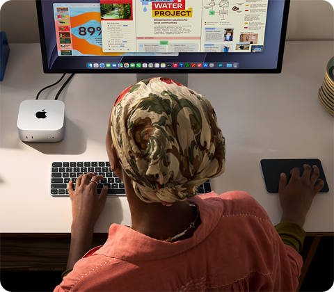 Osoba pisząca przy biurku na Macu mini z podłączonym monitorem Studio Display, klawiaturą Magic Keyboard, gładzikiem Magic Trackpad i różnymi akcesoriami
