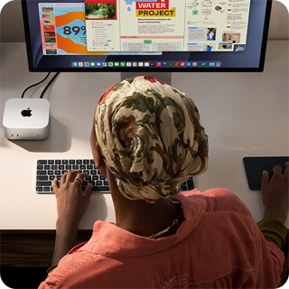 Osoba pisząca przy biurku na Macu mini z podłączonym monitorem Studio Display, klawiaturą Magic Keyboard, gładzikiem Magic Trackpad i różnymi akcesoriami
