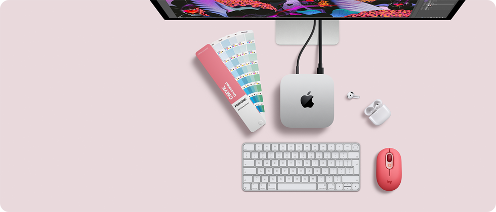 Widoczny z góry Mac mini, aluminium w kolorze srebrnym, zaokrąglone rogi, błyszczące czarne logo Apple pośrodku, podłączony przewodem, częściowo widoczny monitor Studio Display, widoczne słuchawki AirPods 4 w otwartej obudowie, karty kolorów Pantone oraz klawiatura numeryczna i mysz