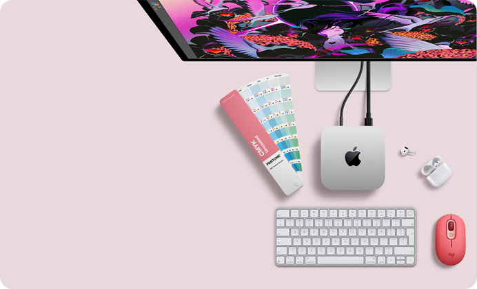 Widoczny z góry Mac mini, aluminium w kolorze srebrnym, zaokrąglone rogi, błyszczące czarne logo Apple pośrodku, podłączony przewodem, częściowo widoczny monitor Studio Display, widoczne słuchawki AirPods 4 w otwartej obudowie, karty kolorów Pantone oraz klawiatura numeryczna i mysz