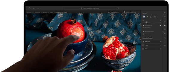 MacBook Pro z wyświetlaczem PROLED XDR, na którym widać martwą naturę z granatami, niebieską ceramiką i tkaninami; dłoń sięga w stronę ekranu, aby wejść w interakcję z obrazem