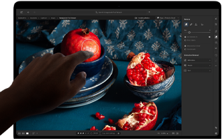 MacBook Pro z wyświetlaczem PROLED XDR, na którym widać martwą naturę z granatami, niebieską ceramiką i tkaninami; dłoń sięga w stronę ekranu, aby wejść w interakcję z obrazem