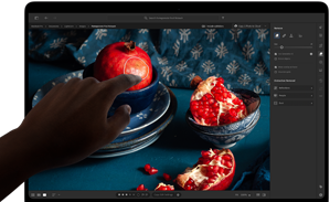 MacBook Pro z wyświetlaczem PROLED XDR, na którym widać martwą naturę z granatami, niebieską ceramiką i tkaninami; dłoń sięga w stronę ekranu, aby wejść w interakcję z obrazem