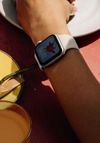 Ręka sięgająca po śniadanie, Apple Watch SE 3 na nadgarstku, koperta z aluminium w kolorze księżycowej poświaty, niegasnący wyświetlacz, portret osoby na tarczy zegarka, pasek sportowy w kolorze księżycowej poświaty