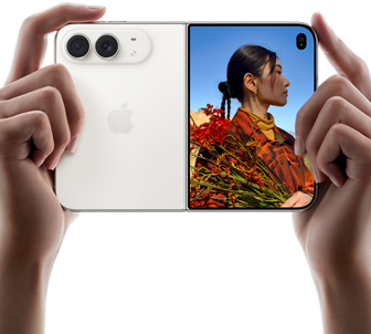 Trzymany w dwóch dłoniach rozłożony iPhone Duo w orientacji poziomej, lewy panel przedstawia aparaty tylne Fusion 48 MP, po prawej stronie na wyświetlaczu zewnętrznym widać osobę pozującą do zdjęcia, widzi ona swoją twarz podczas robienia zdjęcia dzięki funkcji Duo Preview