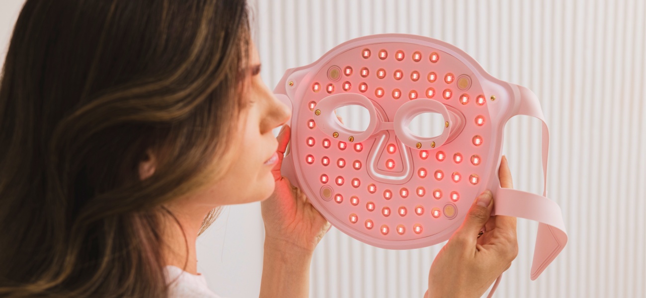 Kobieta trzyma jasnoróżową maskę do terapii światłem LED na twarz Beautifly Aura Glam Pro z podświetlonymi na czerwono diodami, na tle jasnej ściany z pionowymi paskami