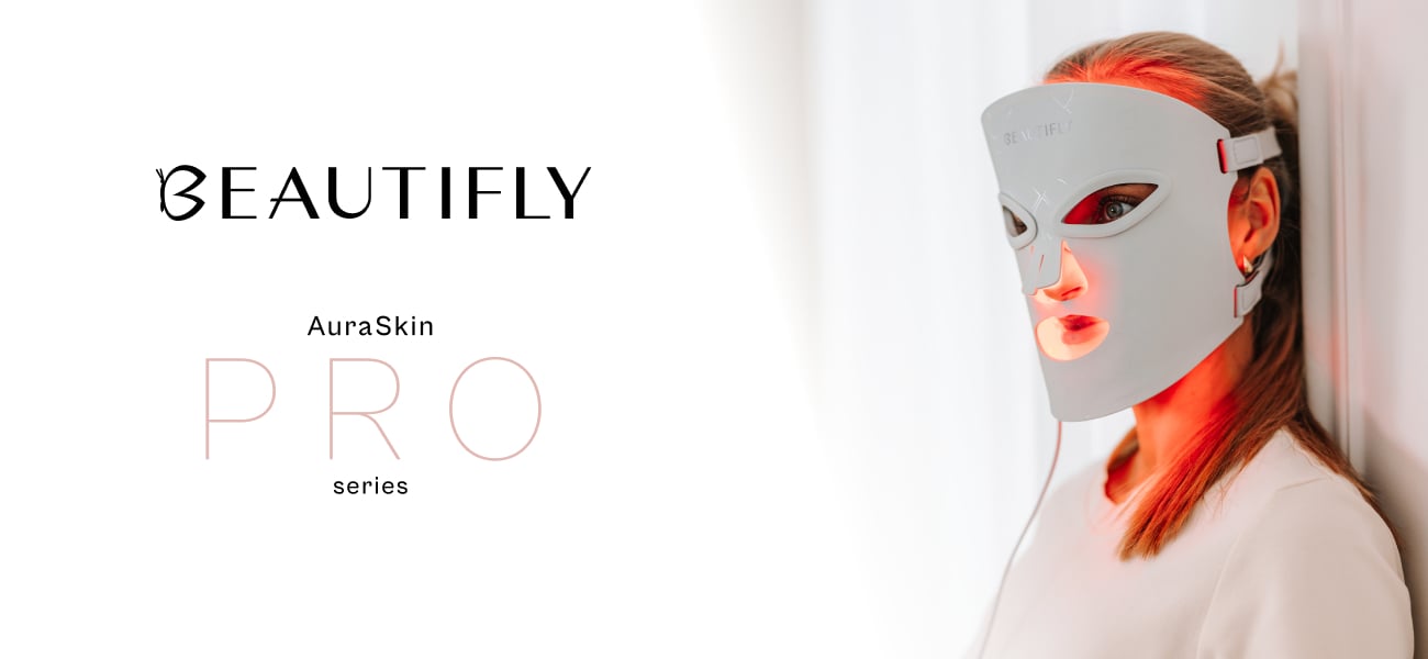 Biała maska LED Beautifly AuraSkin Pro na twarzy kobiety stojącej przy jasnej ścianie, oświetlona czerwonym światłem, na białym tle z czarnym logo marki i nazwą serii PRO