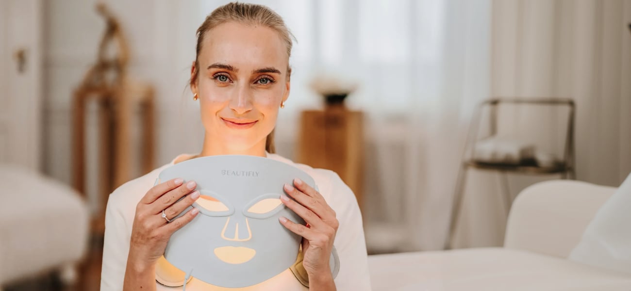 Kobieta w jasnej bluzce trzyma przed sobą białą maskę LED Beautifly AuraSkin Pro z podświetlanymi otworami na oczy i usta, siedząc w jasnym wnętrzu z rozmytym tłem i meblami w beżowych odcieniach