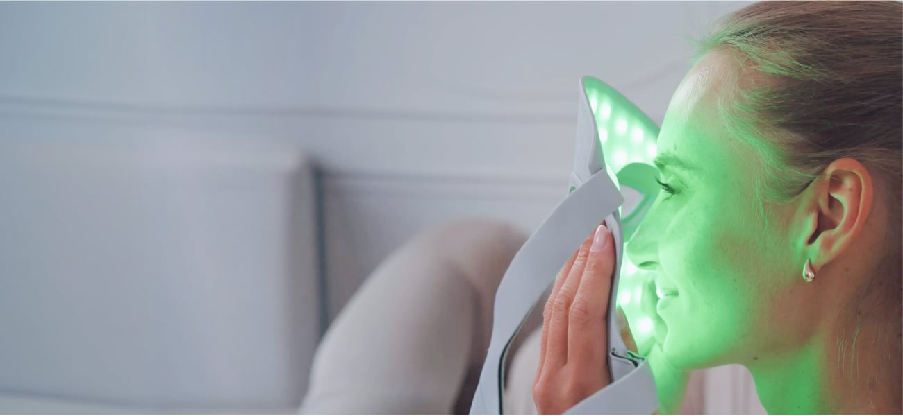 Kobieta przykłada do twarzy białą maskę LED Beautifly AuraSkin Pro emitującą zielone światło, stojąc w jasnym wnętrzu z rozmytym tłem i widocznym profilem twarzy oraz dłonią przytrzymującą urządzenie