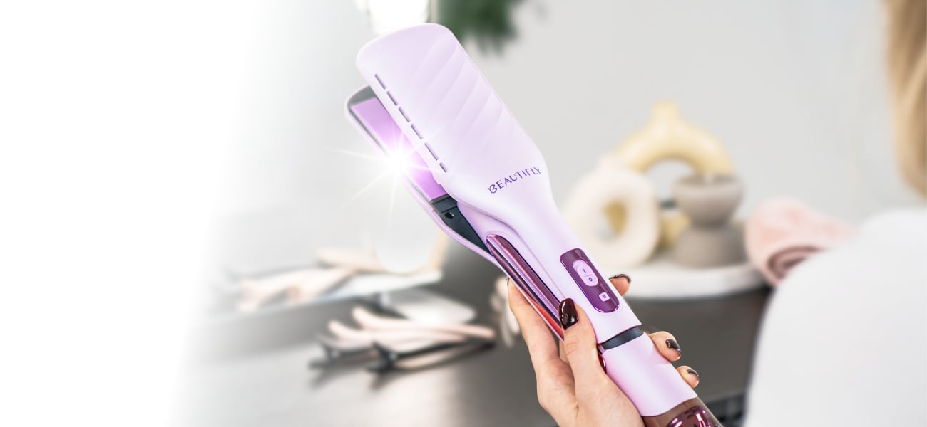 Dłoń trzymająca aeroprostownicę Beautifly DualPerfection z podświetlonym wnętrzem płytek na rozmytym tle toaletki z akcesoriami do włosów, świecą i złożonym ręcznikiem