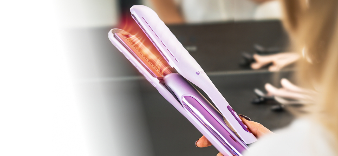 Dłoń trzymająca otwartą aeroprostownicę Beautifly DualPerfection z widocznym pomarańczowym podświetleniem wnętrza urządzenia na rozmytym tle toaletki z akcesoriami do włosów