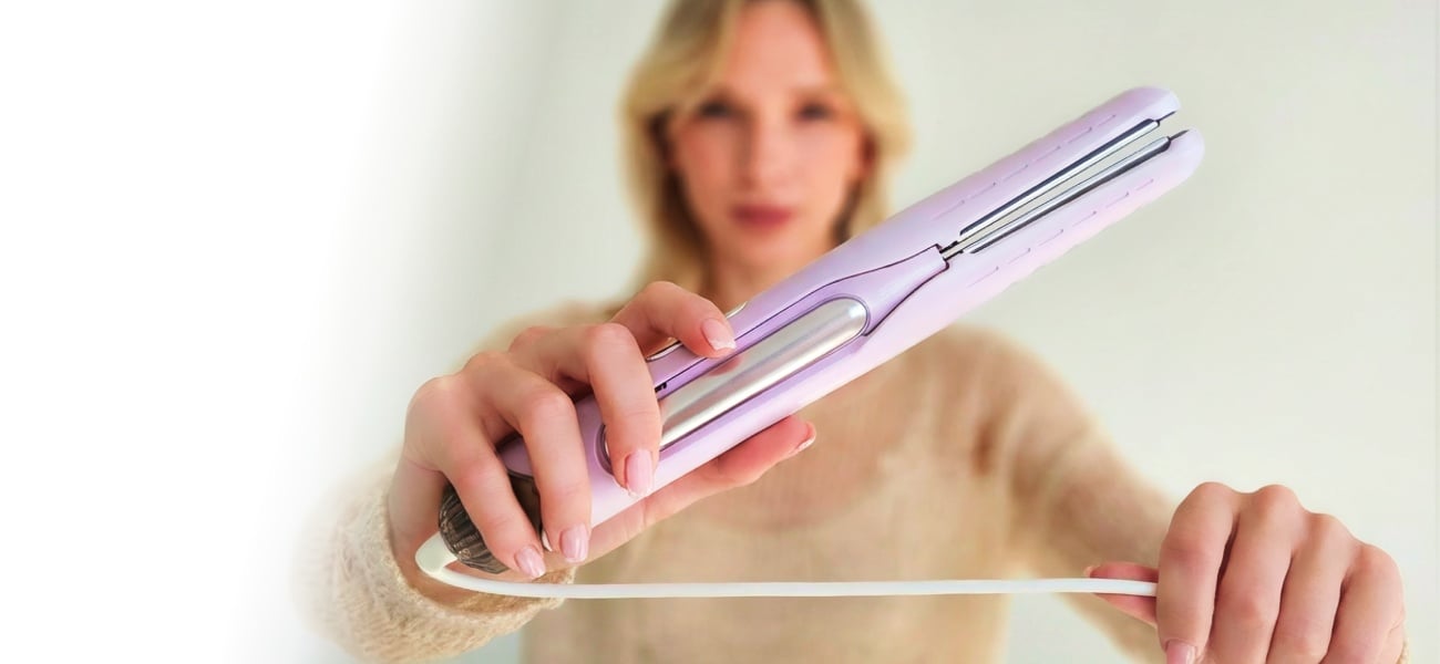Kobieta w jasnym swetrze trzymająca aeroprostownicę Beautifly DualPerfection nad pasmem włosów na jasnym tle, z rozmytą sylwetką w tle i widocznym przewodem urządzenia