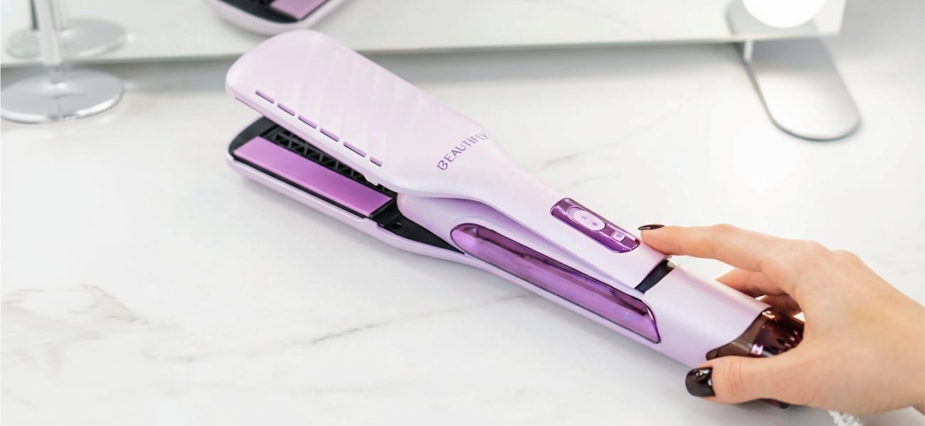 Dłoń dotykająca przycisku otwartej aeroprostownicy Beautifly DualPerfection leżącej na jasnym blacie z widocznymi płytkami prostującymi i rozmytym lustrem w tle