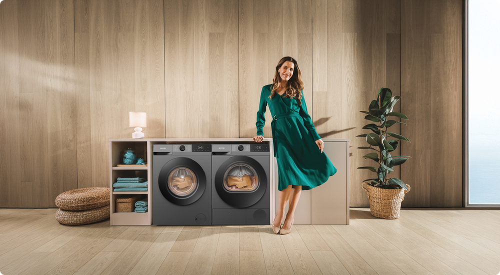 Kobieta w zielonej sukience stoi obok pralki i suszarki marki Gorenje umieszczonych pod blatem