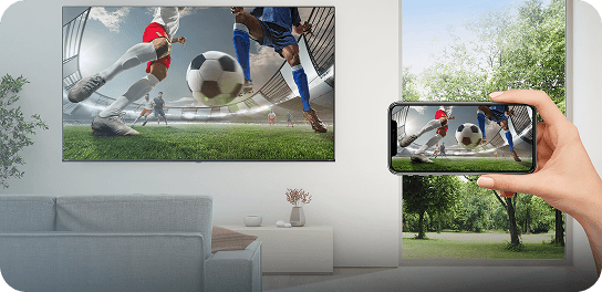 Ręka trzyma smartfon wyświetlający mecz piłki nożnej, przesyłając obraz na ekran telewizora w salonie
