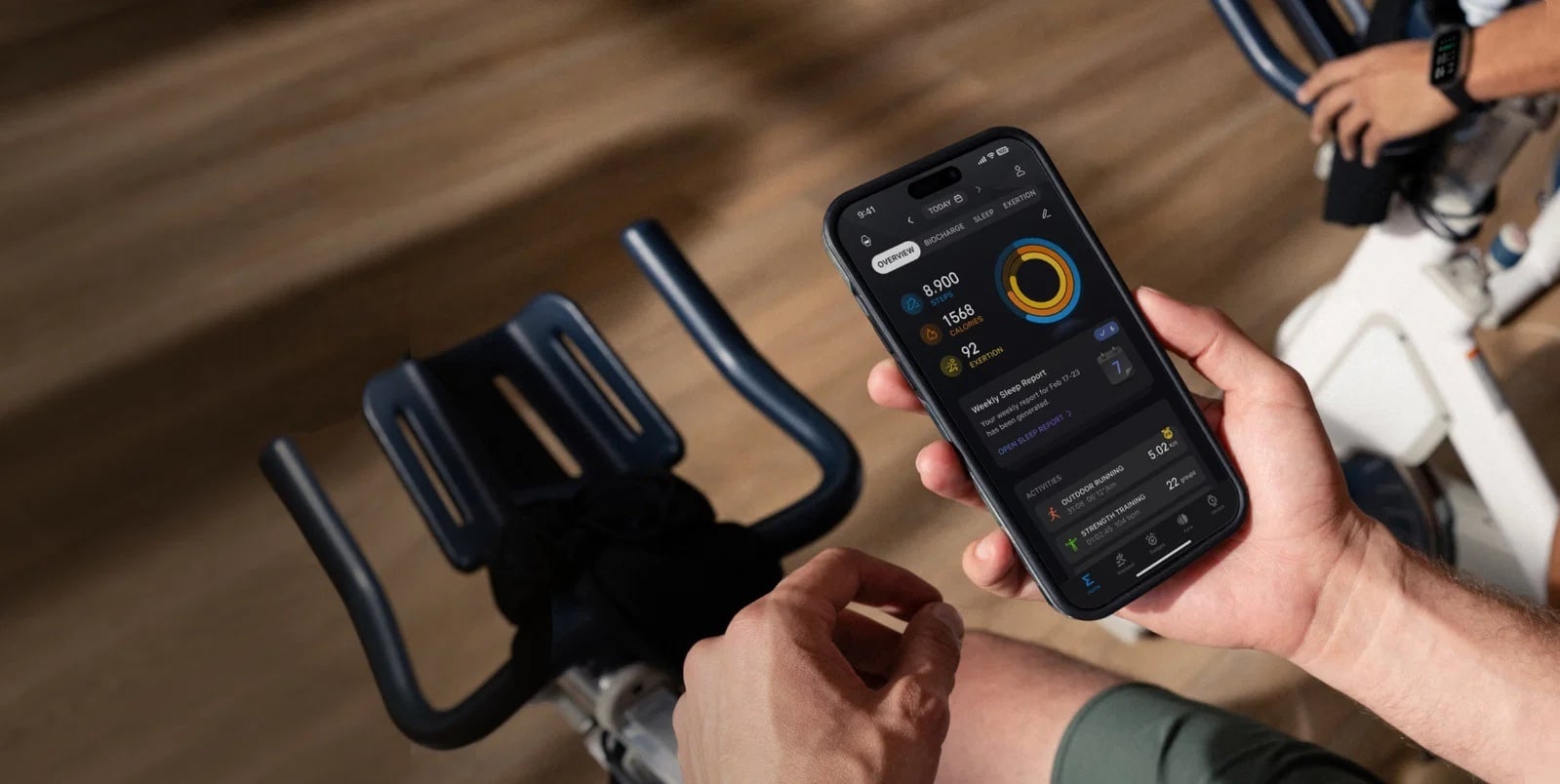 Smartfon z uruchomioną aplikacją fitness wyświetlającą statystyki treningu, trzymany w dłoni na rowerze stacjonarnym.