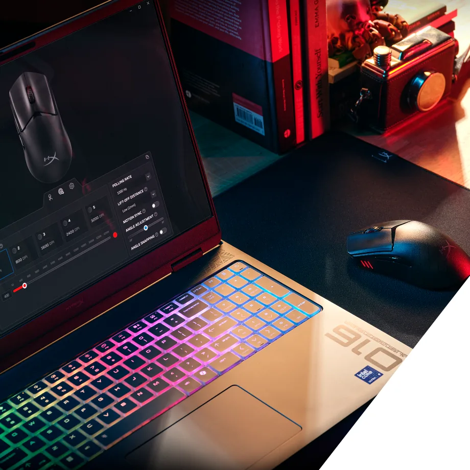 Laptop z podświetlaną klawiaturą i panelem ustawień myszy na ekranie; obok czarna mysz HyperX, książki i aparat