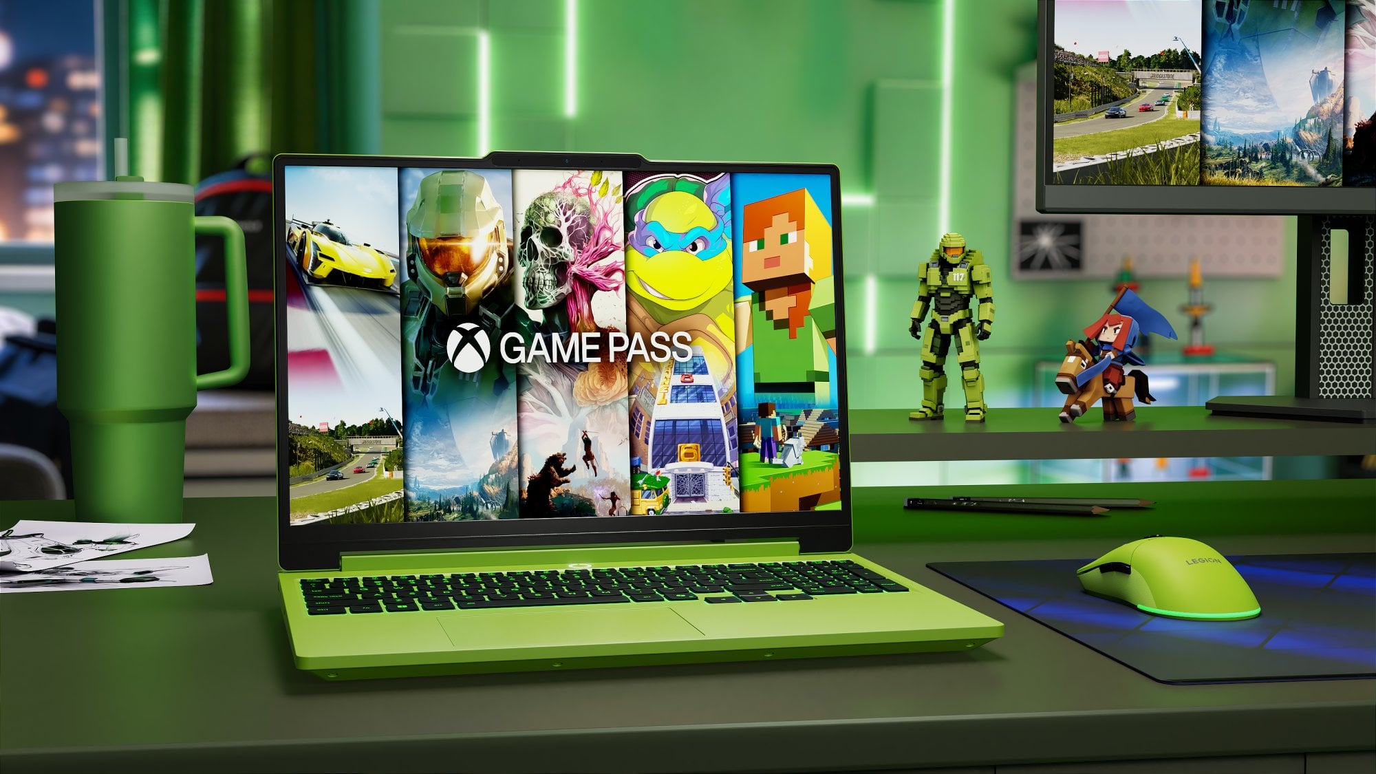 Zielony laptop z otwartą klapą, na ekranie którego wyświetlana jest grafika Xbox Game Pass z fragmentami gier, stoi na biurku obok zielonego kubka termicznego, myszki gamingowej i figurek postaci z gier.