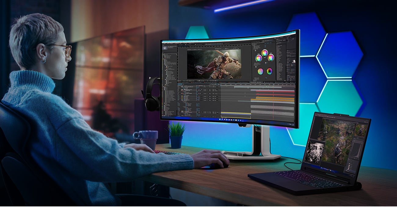 Monitor Lenovo Legion Pro 34WD-10 na biurku przed którym siedzi młoda osoba, obok otwarty laptop podłączony do monitora