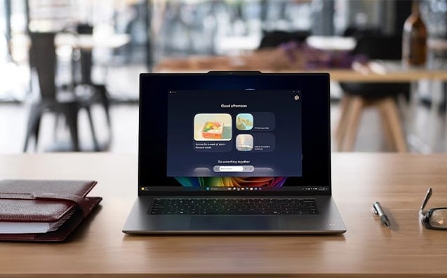 Nowoczesny laptop stoi na drewnianym biurku w kawiarni, na ekranie wyświetla się interfejs z powitaniem "Good afternoon" i propozycjami aktywności, obok leży notatnik i długopis.