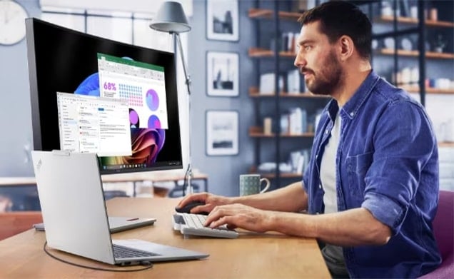 Mężczyzna w niebieskiej koszuli pracuje przy biurku w domu, korzystając z laptopa i zewnętrznego monitora wyświetlającego arkusz kalkulacyjny i wykresy.