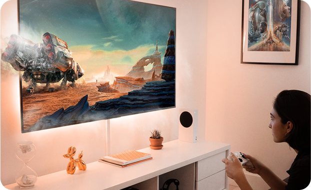 Kobieta siedzi w białym pokoju na wprost ekranu telewizora i gra na konsoli, a duży robot w grze częściowo wychodzi z ekranu