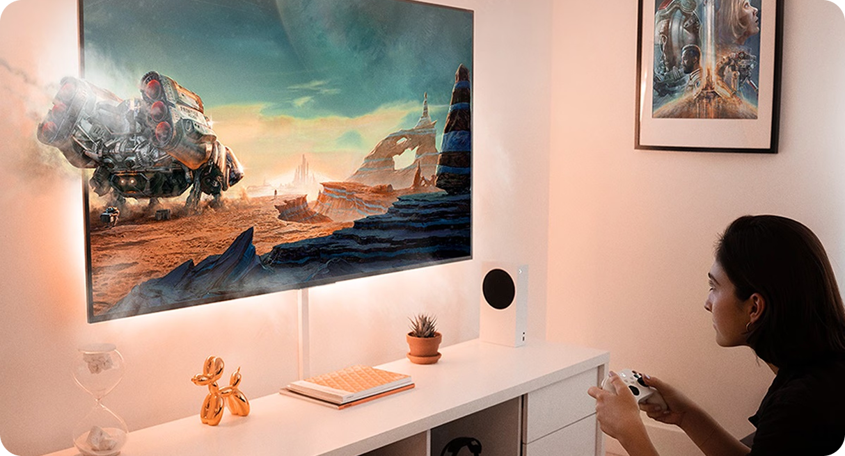 Kobieta siedzi w białym pokoju na wprost ekranu telewizora i gra na konsoli, a duży robot w grze częściowo wychodzi z ekranu