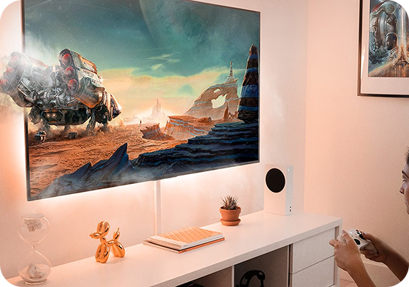 Kobieta siedzi w białym pokoju na wprost ekranu telewizora i gra na konsoli, a duży robot w grze częściowo wychodzi z ekranu