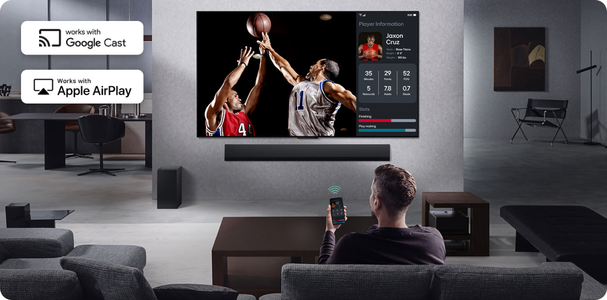 Mężczyzna siedzący na kanapie trzyma w ręce telefon skierowany na ekran telewizora na którym rozgrywa się mecz koszykówki