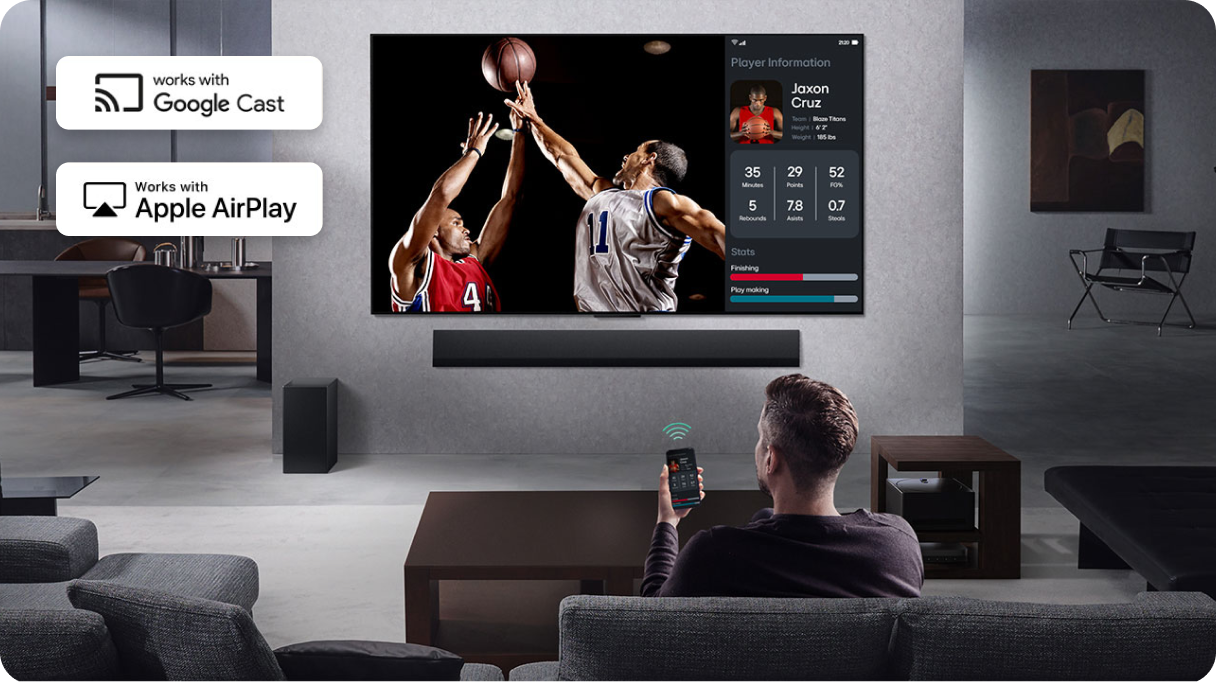 Mężczyzna siedzący na kanapie trzyma w ręce telefon skierowany na ekran telewizora na którym rozgrywa się mecz koszykówki