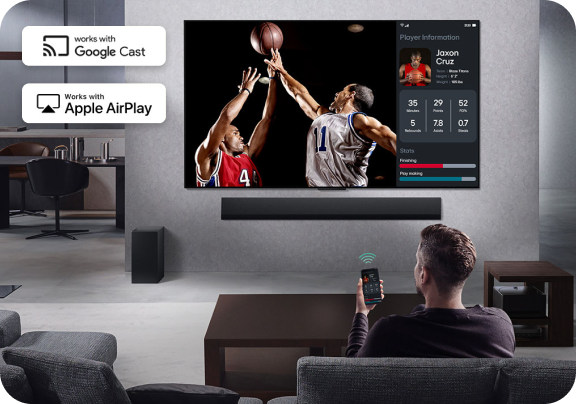 Mężczyzna siedzący na kanapie trzyma w ręce telefon skierowany na ekran telewizora na którym rozgrywa się mecz koszykówki