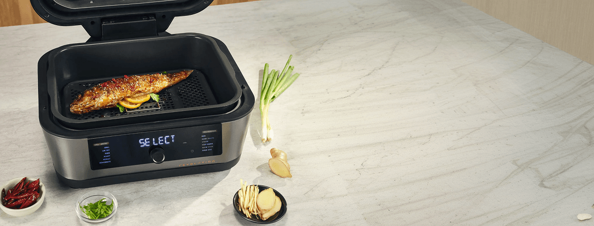 Multicooker z napisem SELECT na panelu, pieczona ryba wewnątrz, obok imbir, szczypiorek i papryczki