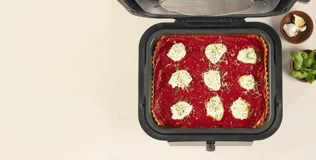 Widok z góry na otwarty multicooker z zapiekaną lasagne w środku, obok którego leży czosnek i świeża bazylia