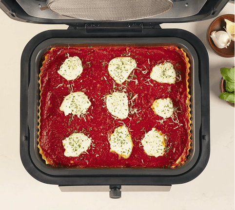 Widok z góry na otwarty multicooker z zapiekaną lasagne w środku, obok którego leży czosnek i świeża bazylia