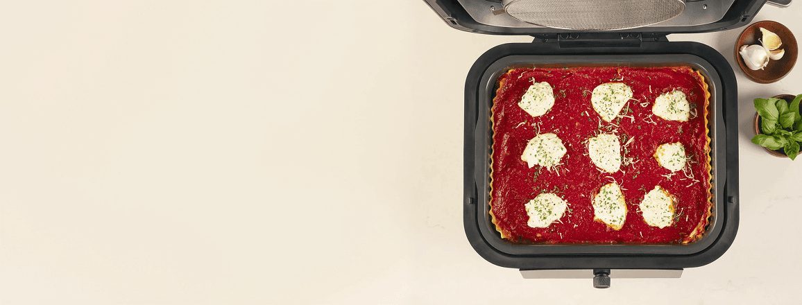 Widok z góry na otwarty multicooker z zapiekaną lasagne w środku, obok którego leży czosnek i świeża bazylia