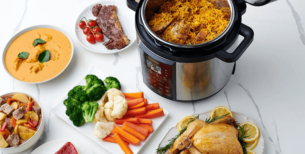 Otwarty multicooker z potrawą z ryżu w środku, otoczony miskami i talerzami z kurczakiem, warzywami oraz gulaszem
