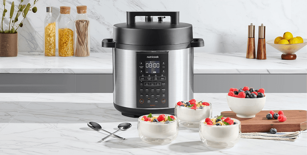 Srebrny multicooker z czarną pokrywą stojący na blacie kuchennym obok trzech szklanych salzerek z deserem z malinami