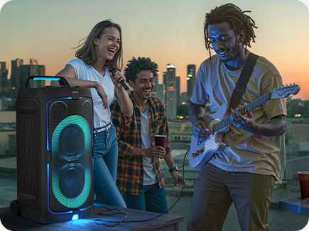 data-desktop-alt="3 people outdoors are enjoying themselves. One person plays the guitar and one sings via microphone with the Sound Tower."