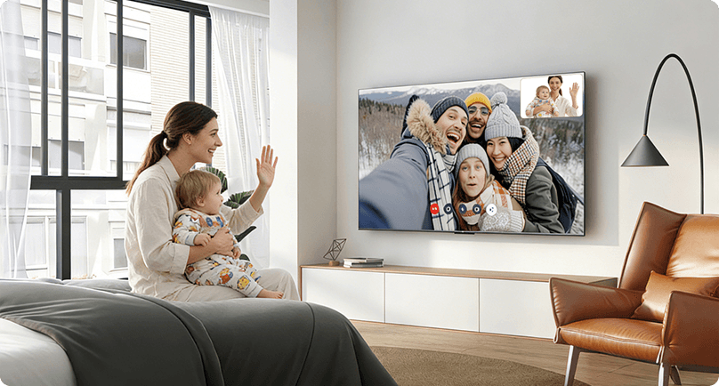 Kobieta z dzieckiem machająca do telewizora TCL A400 wyświetlającego wideorozmowę z grupą uśmiechniętych osób.