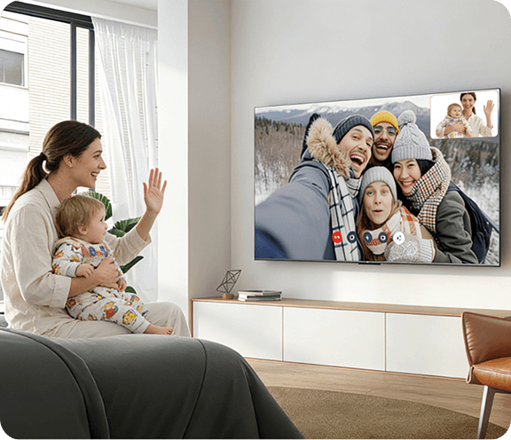 Kobieta z dzieckiem machająca do telewizora TCL A400 wyświetlającego wideorozmowę z grupą uśmiechniętych osób.