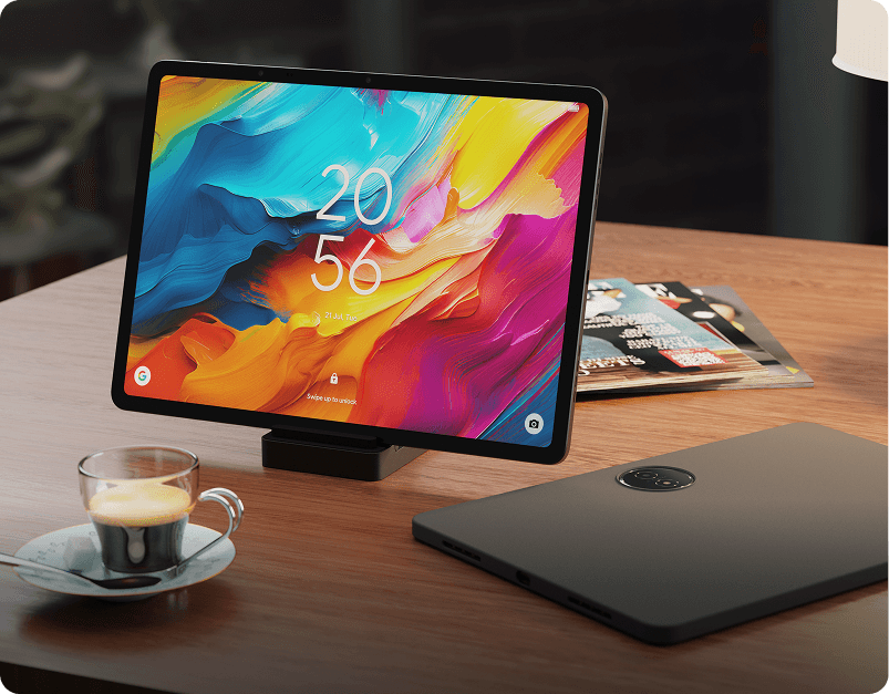 Tablet w stacji dokującej z kolorową tapetą stojący na biurku obok filiżanki kawy.