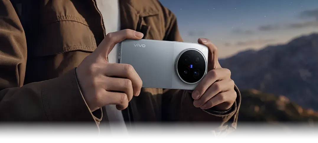 Osoba w brązowej kurtce trzyma poziomo srebrny smartfon vivo z aparatami ZEISS na tle gór