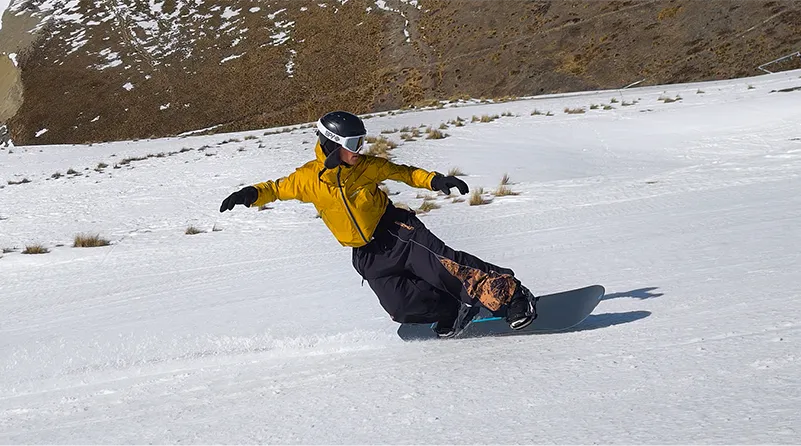 Snowboardzista w żółtej kurtce i czarnym kasku skręca na ośnieżonym górskim stoku