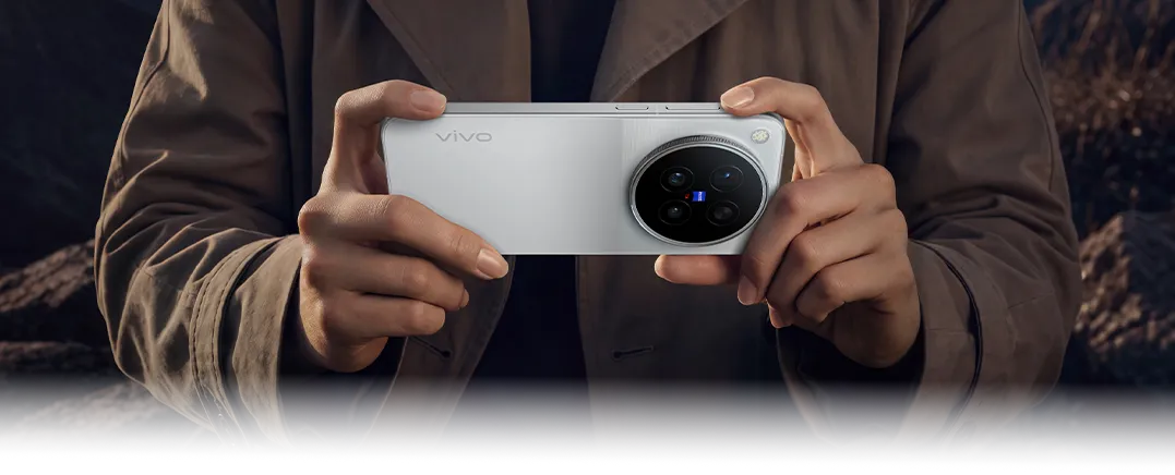Osoba w brązowej kurtce trzyma poziomo srebrny smartfon vivo z aparatami ZEISS na tle gór